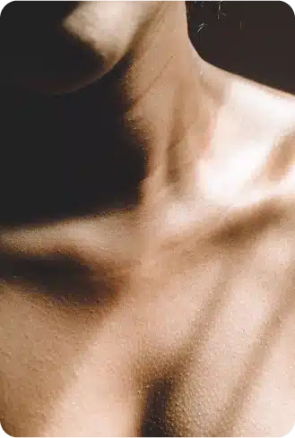 Nahaufnahme des Halses, des Schlüsselbeins und des oberen Brustbereichs einer Person, mit weicher Beleuchtung und Schatten, die die Konturen der Haut hervorheben - ideal, um subtile Ergebnisse nach einer Brustvergrößerung zu zeigen.