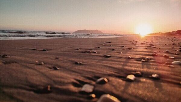 Ein Sandstrand bei Sonnenuntergang, aus einem niedrigen Winkel betrachtet, ruft Ruhe hervor. Kleine Kieselsteine bedecken den Sand, während sich sanfte Wellen dem Ufer nähern und die Sonne in der Nähe der fernen Berge untergeht - eine Gelassenheit, die es mit der Ruhe nach Fettabsaugung aufnehmen kann.