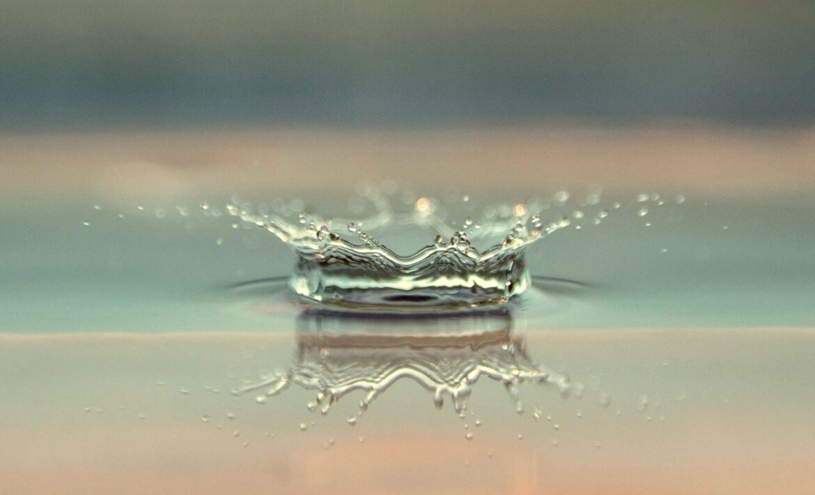Eine Nahaufnahme eines Wassertropfens, der auf eine ruhige Oberfläche trifft und einen kronenförmigen Spritzer mit Wellen und winzigen Tröpfchen erzeugt - ähnlich der Präzision, die man in Fettabsaugung sieht -, der sich auf dem glatten Wasser darunter spiegelt. Der Hintergrund ist mit gedeckten Farben sanft verschwommen.