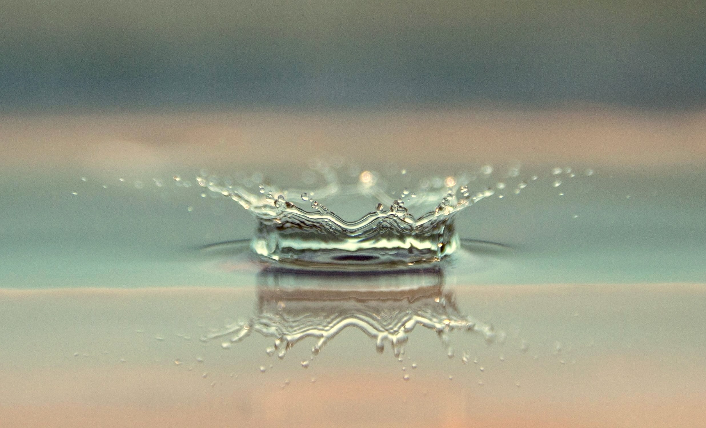 Eine Nahaufnahme eines Wassertropfens, der auf eine ruhige Oberfläche trifft und einen kronenförmigen Spritzer mit Wellen und winzigen Tröpfchen erzeugt - ähnlich der Präzision, die man in Fettabsaugung sieht -, der sich auf dem glatten Wasser darunter spiegelt. Der Hintergrund ist mit gedeckten Farben sanft verschwommen.