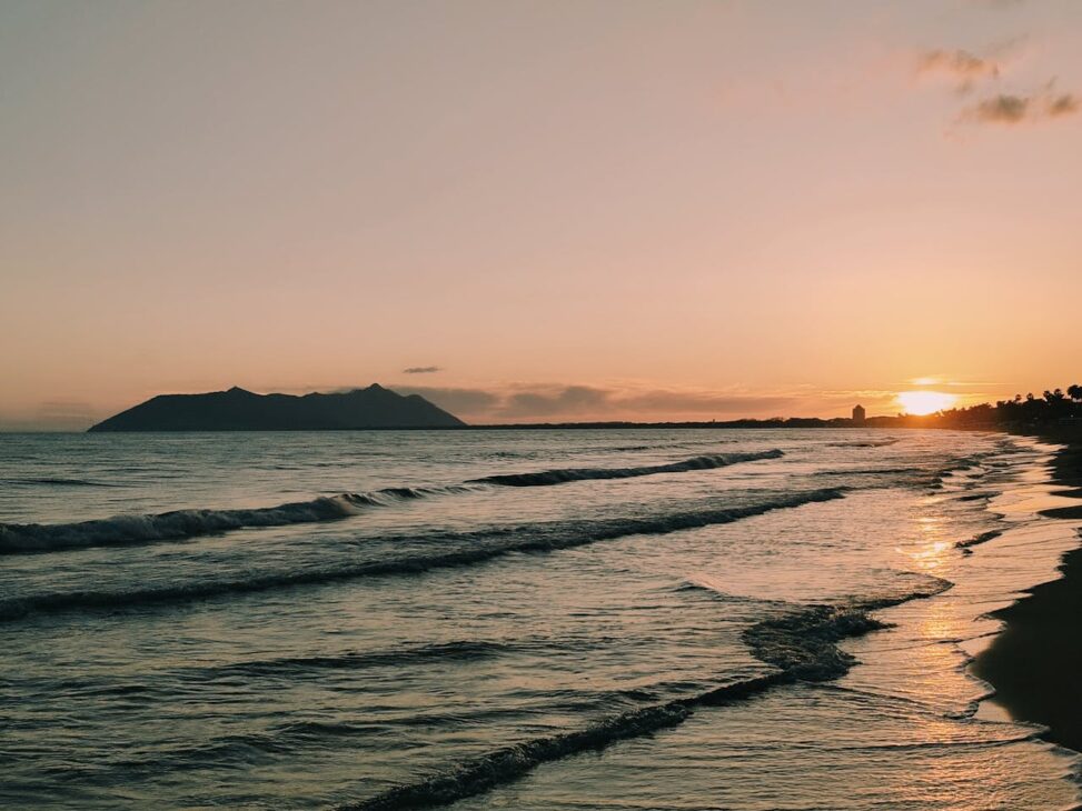 Eine ruhige Strandszene bei Sonnenuntergang mit sanften Wellen, einer silhouettierten Insel in der Ferne und einer glühenden Sonne tief am Horizont, ähnlich wie die ruhige Zuversicht nach Brustkorrekturen in Nürnberg.