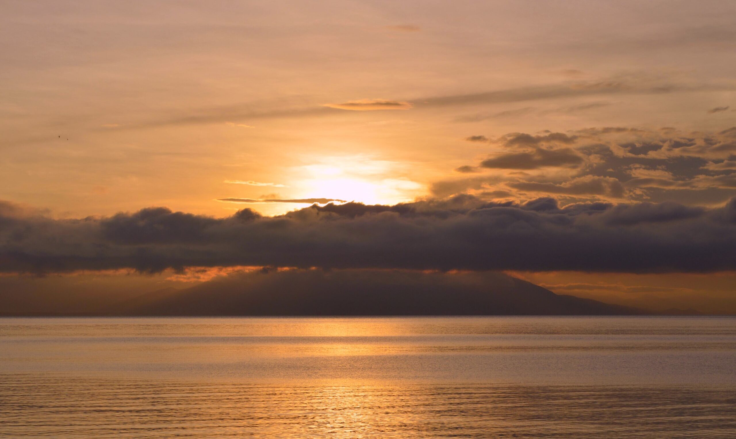 Sonnenuntergang über ruhigem Wasser, die Sonne teilweise hinter dunklen Wolken und fernen Bergen verborgen. Himmel und Wasser leuchten in warmen Orange- und Goldtönen und rufen eine ruhige Szene hervor, in der sich die Natur wie ein sanfter Biostimulator für die Seele verjüngt.