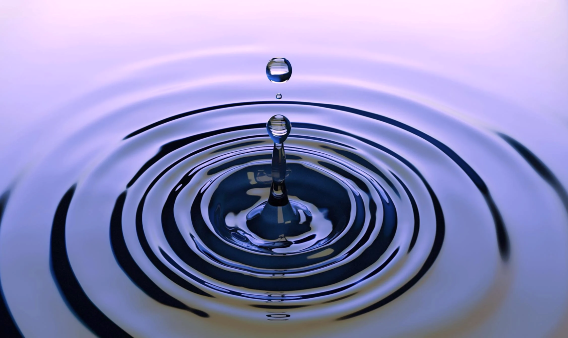 Eine Nahaufnahme eines Wassertropfens, der auf eine glatte Wasseroberfläche fällt und konzentrische Wellen mit sanften Reflexen erzeugt - ähnlich wie die Präzision der Fettabsaugung. Die Farben sind Schattierungen von Blau und Violett.