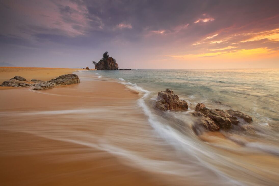 Goldener Sandstrand mit sanften Wellen, die die Felsen umspülen, eine kleine Felseninsel mit Bäumen in der Ferne, unter einem dramatischen, farbenfrohen Sonnenuntergangshimmel - perfekt zum Entspannen, nachdem Sie Möglichkeiten wie Brustkorrekturen in Nürnberg erkundet haben.